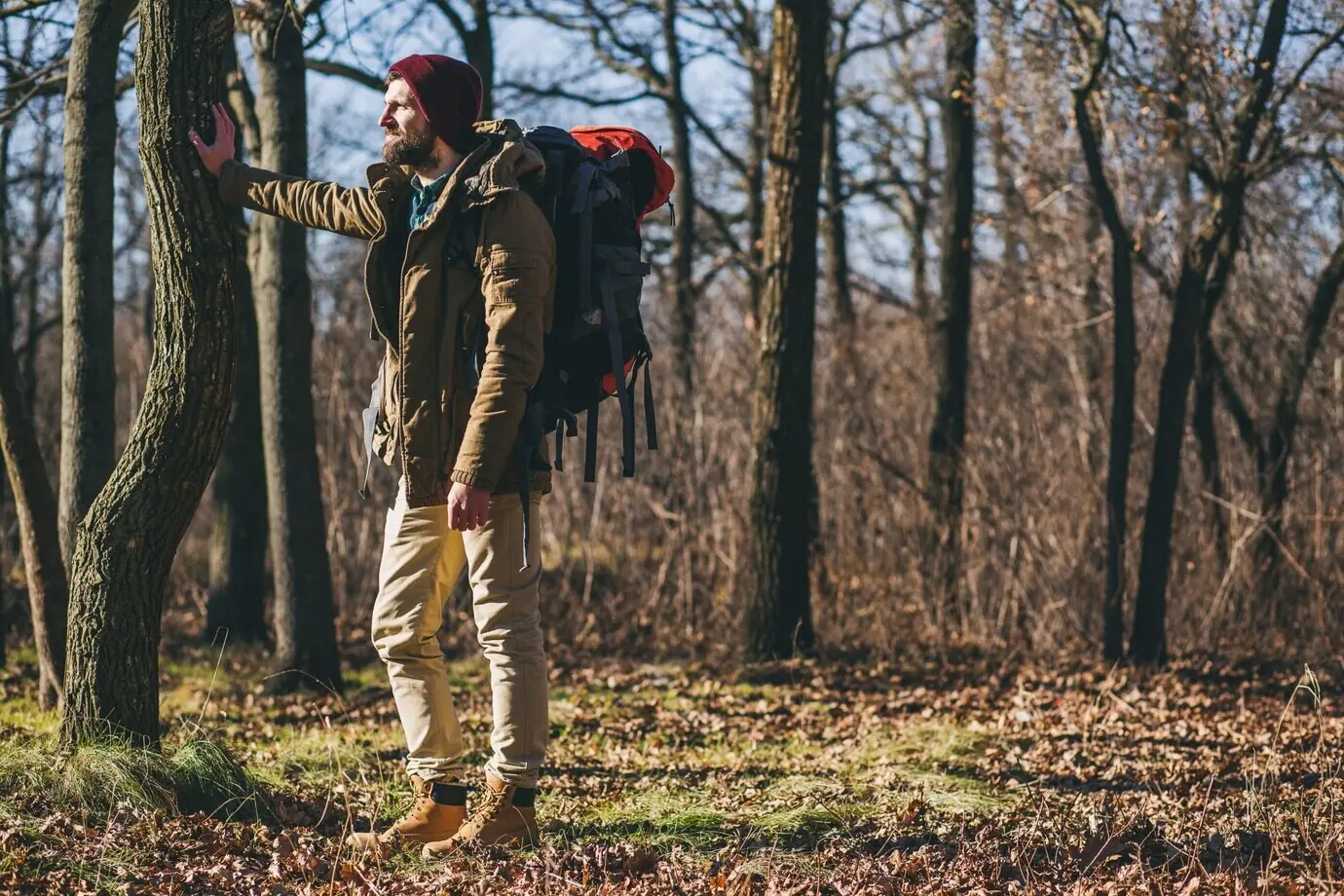 Hipster-Mann mit Rucksack unterwegs in einem herbstlichen Wald, trägt warme Jacke und Mütze; aktiver Tourist, der in der kalten Jahreszeit die Natur erkundet.